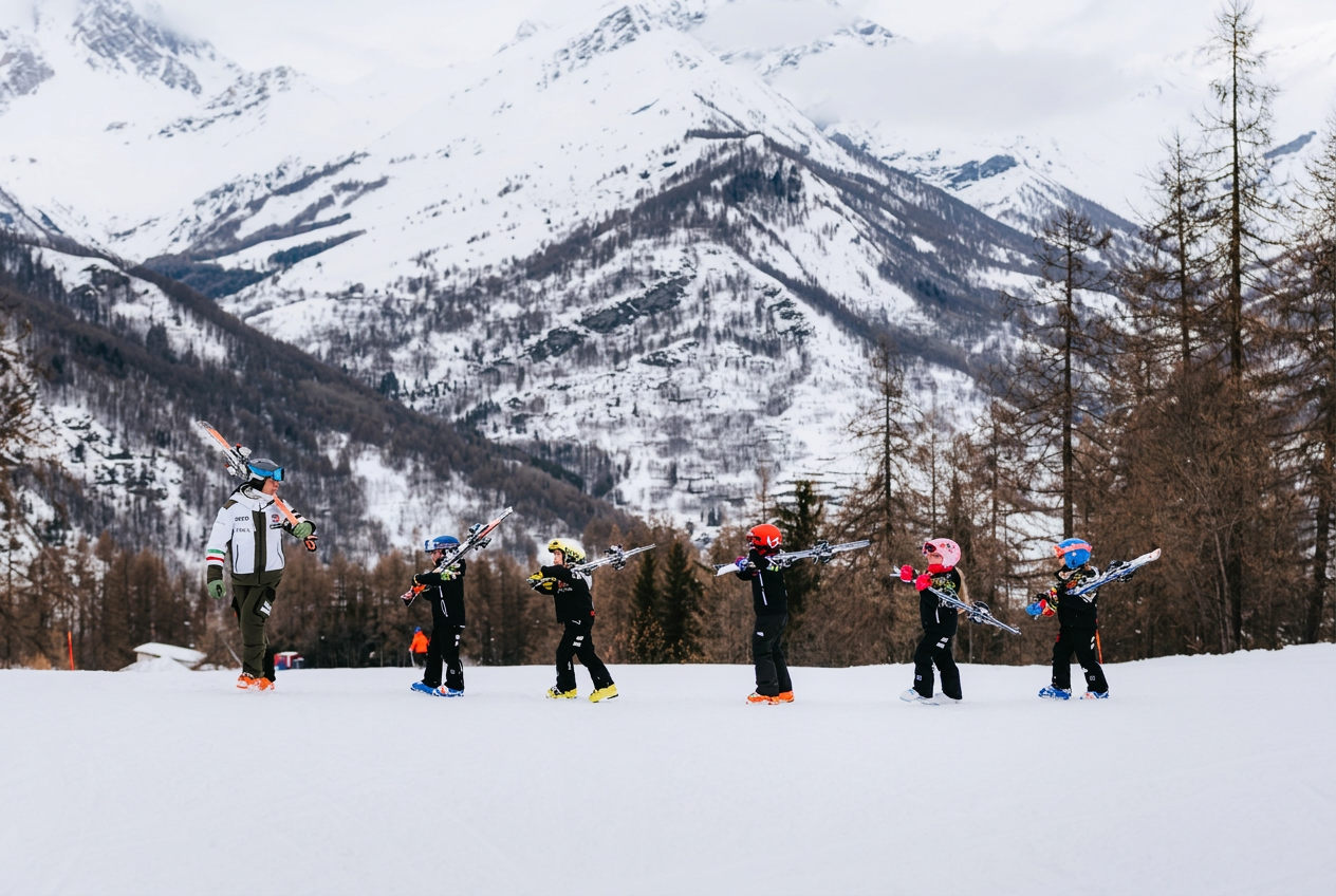 Un moniteur de ski guide un groupe de jeunes enfants portant leurs skis sur une pente enneigée, avec les montagnes en arrière-plan, lors de cours de ski pour enfants destinés aux skieurs expérimentés de l'école de ski Liberi Tutti.