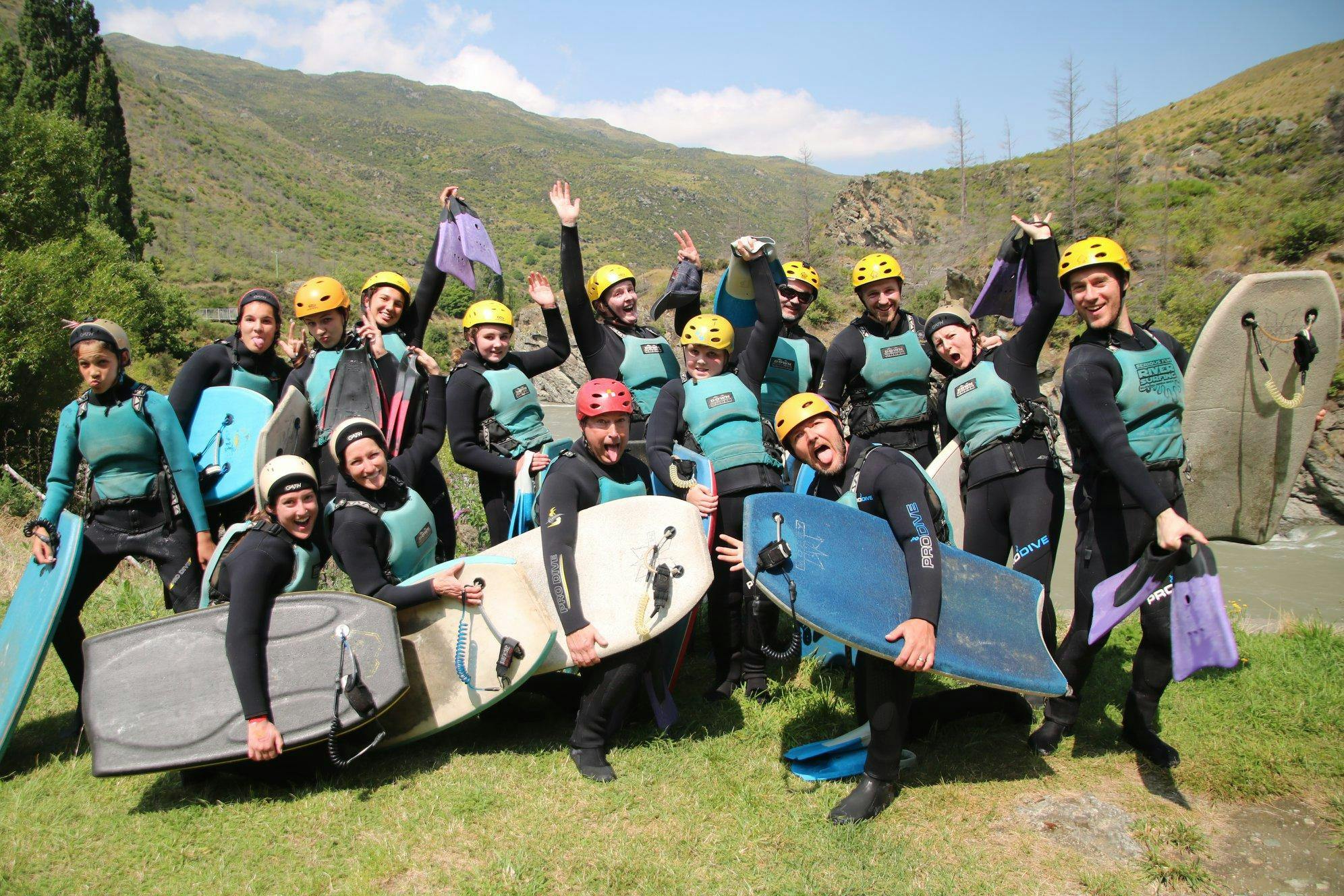 A family is smiling at the camera before jumping into the waters of the Kawarau River to enjoy the activity Riversurfing on the Kawarau River for Families organized by Serious Fun Riverboarding.