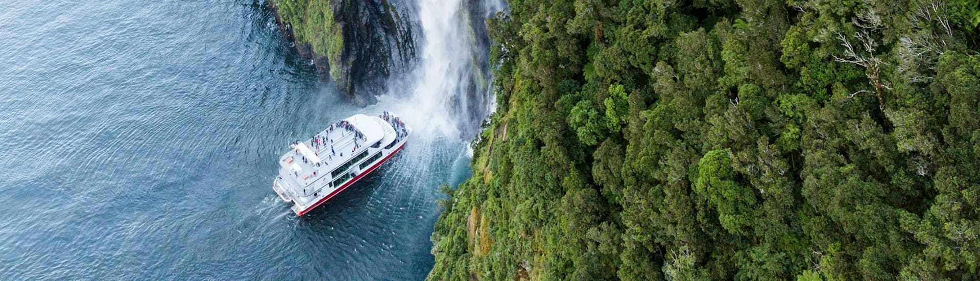 A catamaran operated by Southern Discoveries is navigating towards a majestic waterfall so that the people on the Milford Sound Scenic Day Trip from Te Anau can feel the spray of the water.