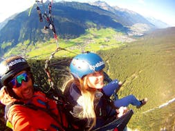 A girl flies together with her tandem pilot from Fly-Stubai over the beautiful mountain world and smiles happily into the camera during the offer "Extended High Altitude Tandem Paragliding in Stubaital".