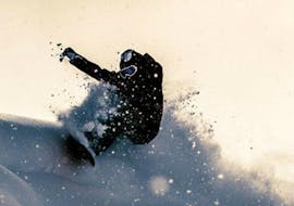 Un snowboardeur fait un saut et fait voler la neige pendant les cours de snowboard freeride - Tous niveaux de l'école de snowboard BOARD.AT.
