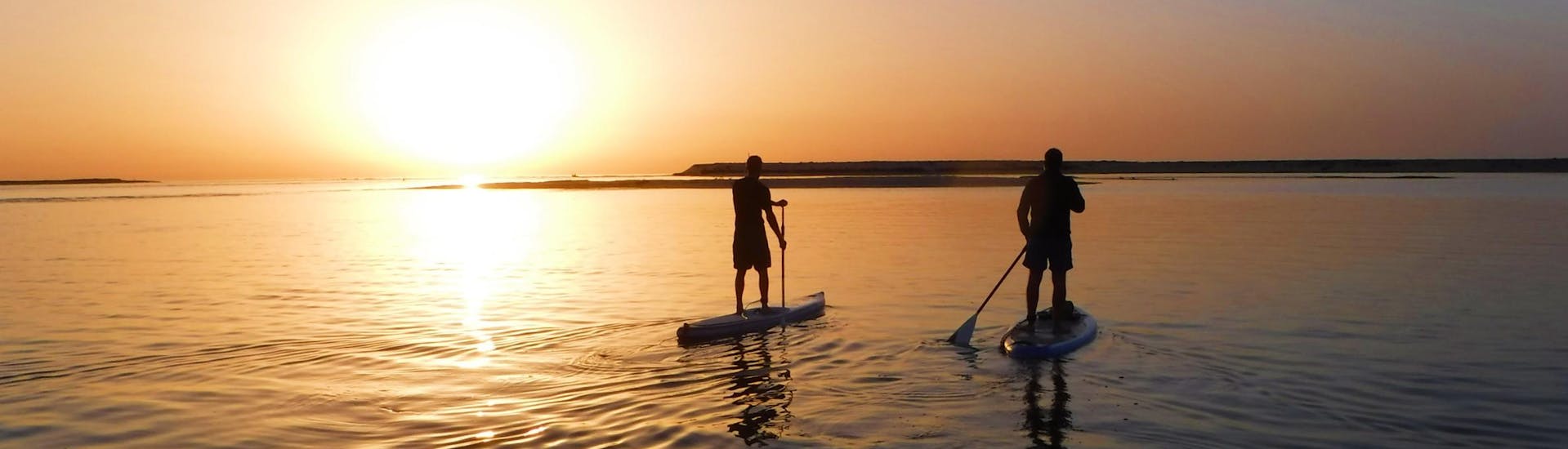 Während des SUP Yoga an der Praia da Fuseta bei Sonnenuntergang mit Kite Culture Algarve paddeln zwei Freunde in den wunderschönen Sonnenuntergang über der Lagune.