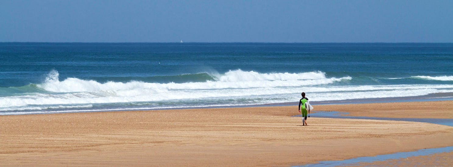 A young surfer is walking along the beach where surfing lessons are taking place with Capbreton Surfer School.