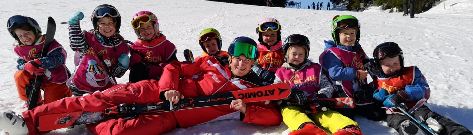 Eine Gruppe kleiner Skifahrer mit ihrem Skilehrer liegen im Schnee während ihrem Kinder-Skikurs Bambini mit der Skischule Reiteralm.