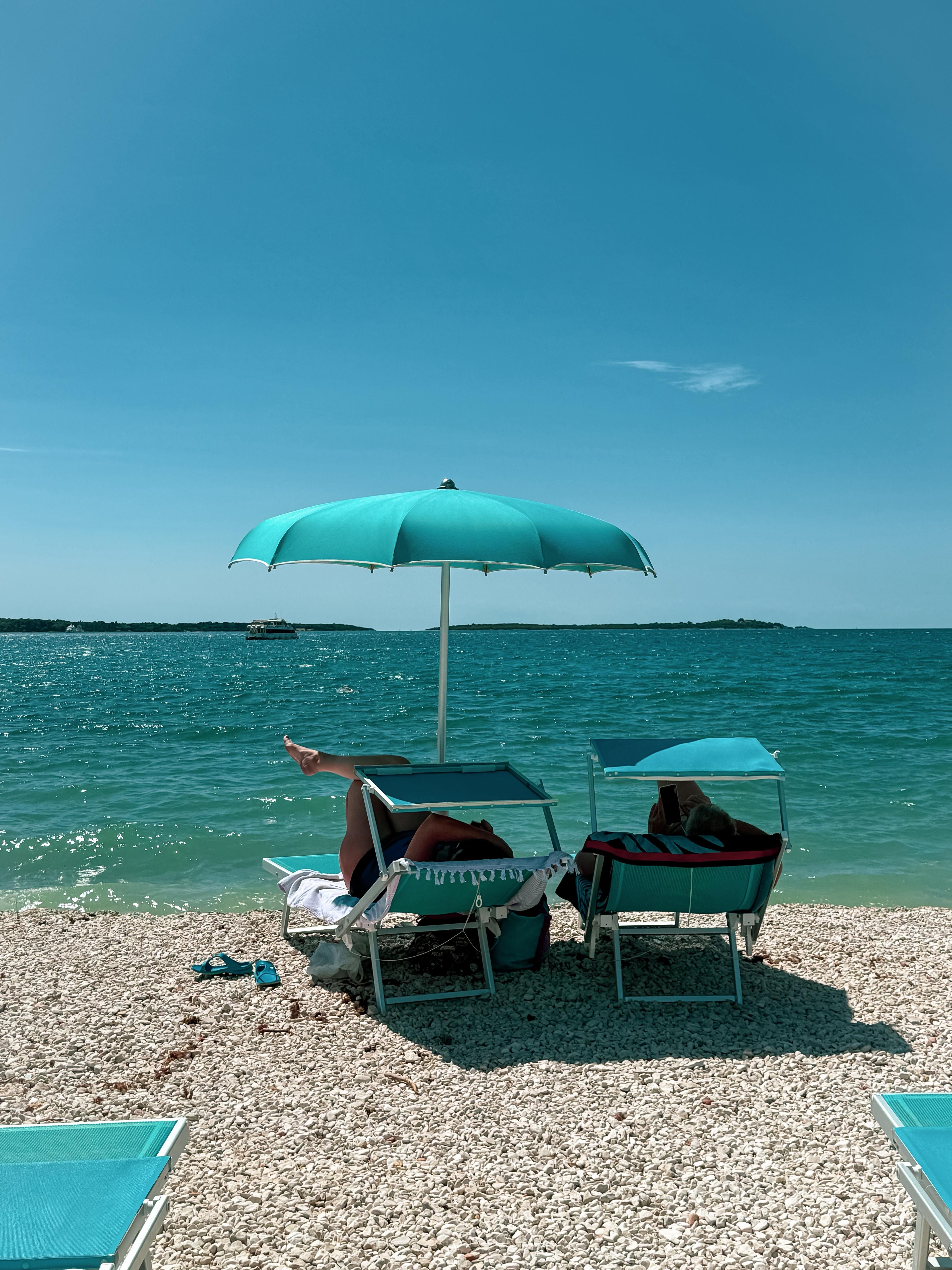 Twee mensen ontspannen op ligbedden onder een turquoise parasol op een kiezelstrand tijdens een boottocht rond het Brijuni Nationaal Park in Pula, inclusief lunch verzorgd door Pula Boat Tours-Adventures.