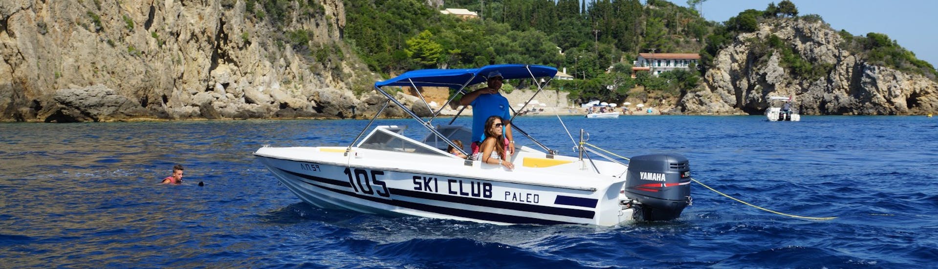 Un bateau sur la mer et une personne faisant de la planche à voile dans l'eau pendant la session de planche à voile à la plage de St. Petros à Corfou avec le Ski Club 105.