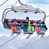 Ein Skilehrer sitzt im Sessellift mit 5 kleinen Skifahrern beim Kinder-Skikurs für Fortgeschrittene.