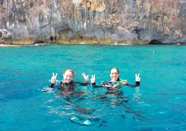 Twee meisjes in het turquoise water met wetsuits, lachend naar de camera tijdens de snorkeltrip voor de kust van Santa Maria Navarrese.