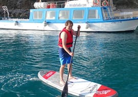 A participant has fun paddling in front of a boat through the Bay of Alcúdia during a SUP board rental in Alcanada Beach with North Coast Adventure Mallorca.