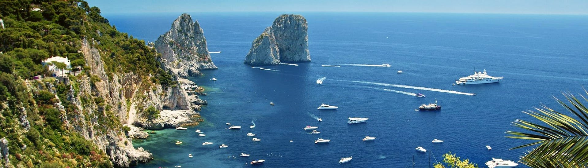 Vista dei faraglioni di Capri dall'alto e delle barche che ci navigano vicino, tra cui quella di HP Travel durante la Gita in barca da Sorrento a Capri con degustazione di limoncello.