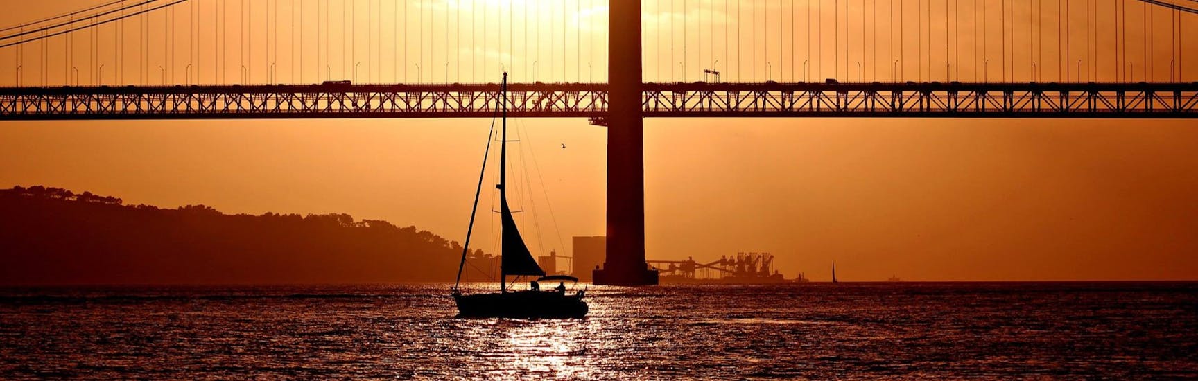 A beautiful sunset with the 25 de Abril Bridge on the background, and a sailboat with participants having fun during a sailing boat trip with Taguscruises Lisbon.