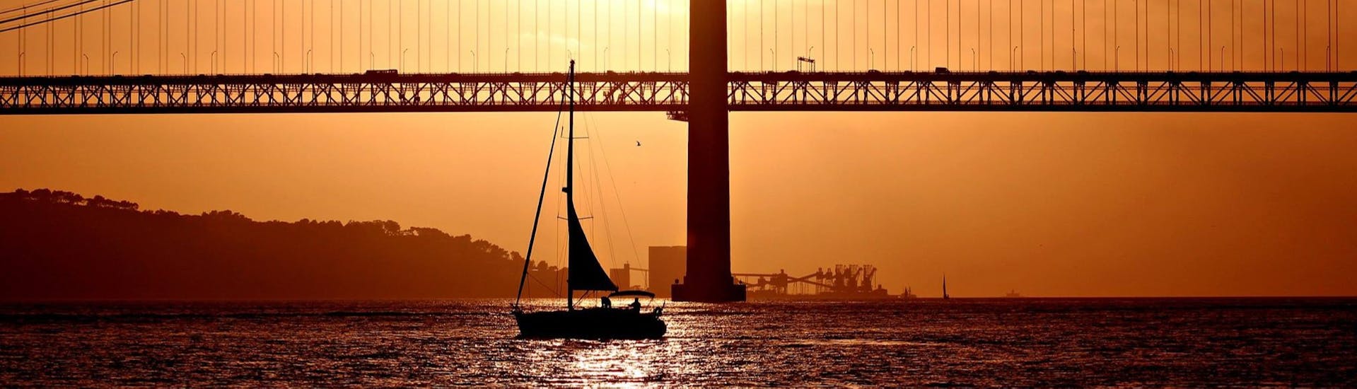 A beautiful sunset with the 25 de Abril Bridge on the background, and a sailboat with participants having fun during a sailing boat trip with Taguscruises Lisbon.