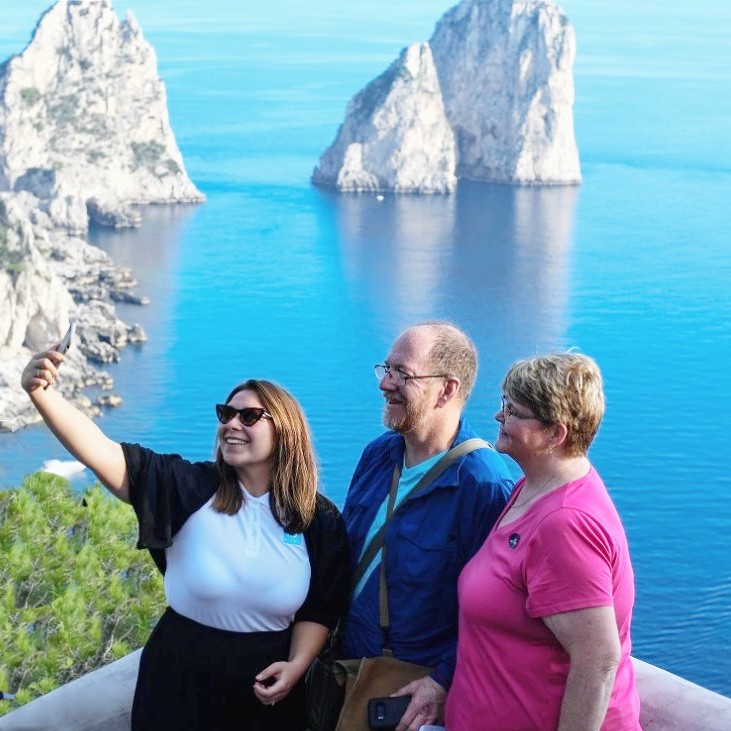 Tres personas tomándose un selfie con el fondo de los Faraglioni durante la excursión en barco por Capri con visita guiada a la isla con HP Travel Capri.