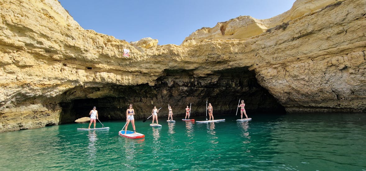 Eine Gruppe von Leuten kommt während der geführten Stand-Up-Paddling-Tour von Praia de Albandeira zur Benagil-Höhle mit Surf4Fun Albufeira auf ihren SUP-Boards aus einer Höhle heraus.