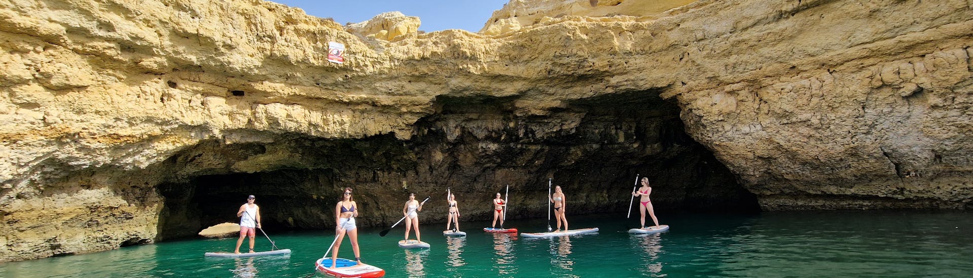 Eine Gruppe von Leuten kommt während der geführten Stand-Up-Paddling-Tour von Praia de Albandeira zur Benagil-Höhle mit Surf4Fun Albufeira auf ihren SUP-Boards aus einer Höhle heraus.