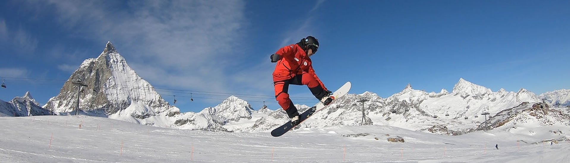 A snowboard instructor jumps over a ramp in front of the mountain panorama of Zermatt during Private Snowboarding Lessons for Kids & Adults of All Levels with the Evolution Ski School Zermatt.