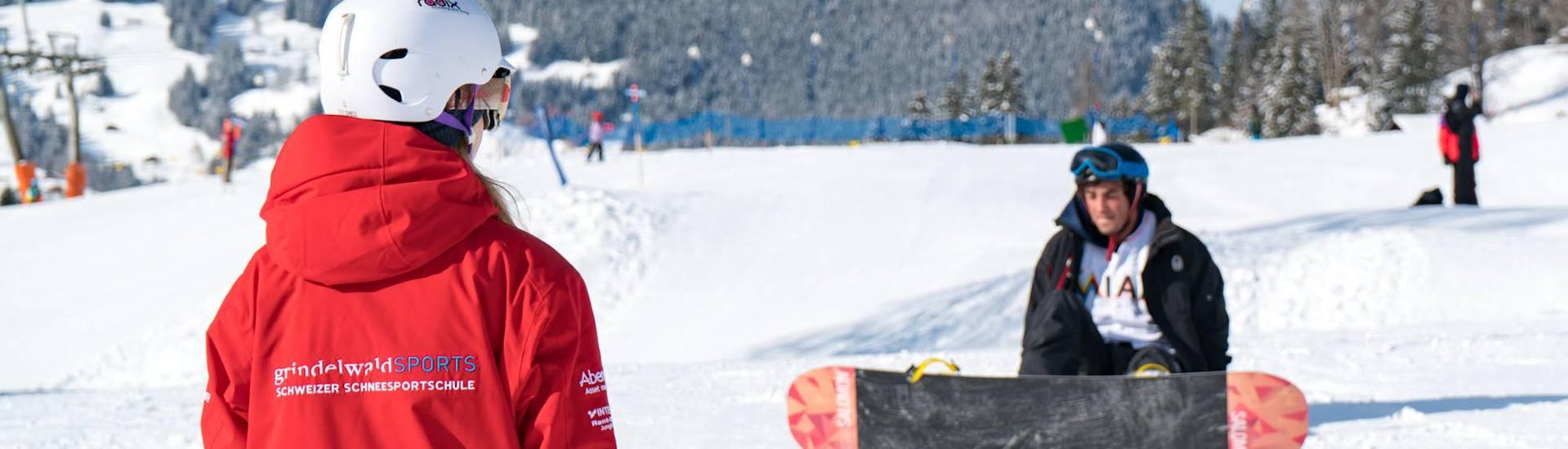 Eine Snowboardlehrerin zeigt einem Teilnehmer wie man richtig aufsteht beim Snowboardkurs + Snowboardverleih Package für Erwachsene für Anfänger mit der Swiss Ski School Grindelwald.