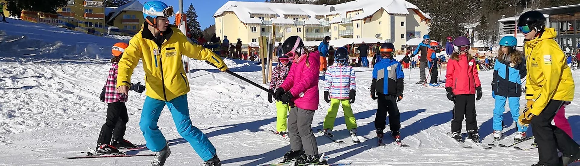 A ski instructor pulls a child with the poles and shows her the right form during the kids ski lessons (4-13 yrs) for all levels with Schneesportschule ON SNOW Feldberg.