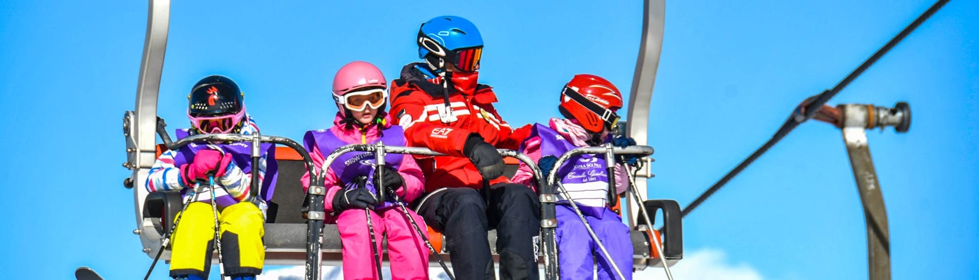 An instructor from Scuola di Sci Pila is sitting on a chairlift with some kids during the Kids Ski Lessons (5-12 y.) for All Levels.