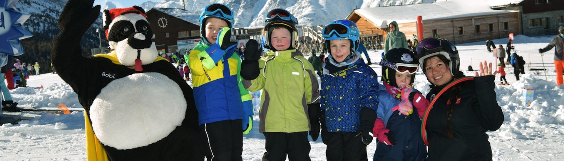 Een groep skiërs gaat op de foto met de mascotte Bobi na een succesvolle cursus bij Bobi's Snowkindergarden (2,5-4 jaar) voor beginners bij de skischool Grächen - Zenklusen Sport.