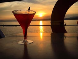 Cocktail rosso su un tavolo con un bellissimo tramonto della Calcidica durante il giro in barca dell'isola di Ammouliani con le crociere giornaliere e private di Eirinikos Glassbottom.