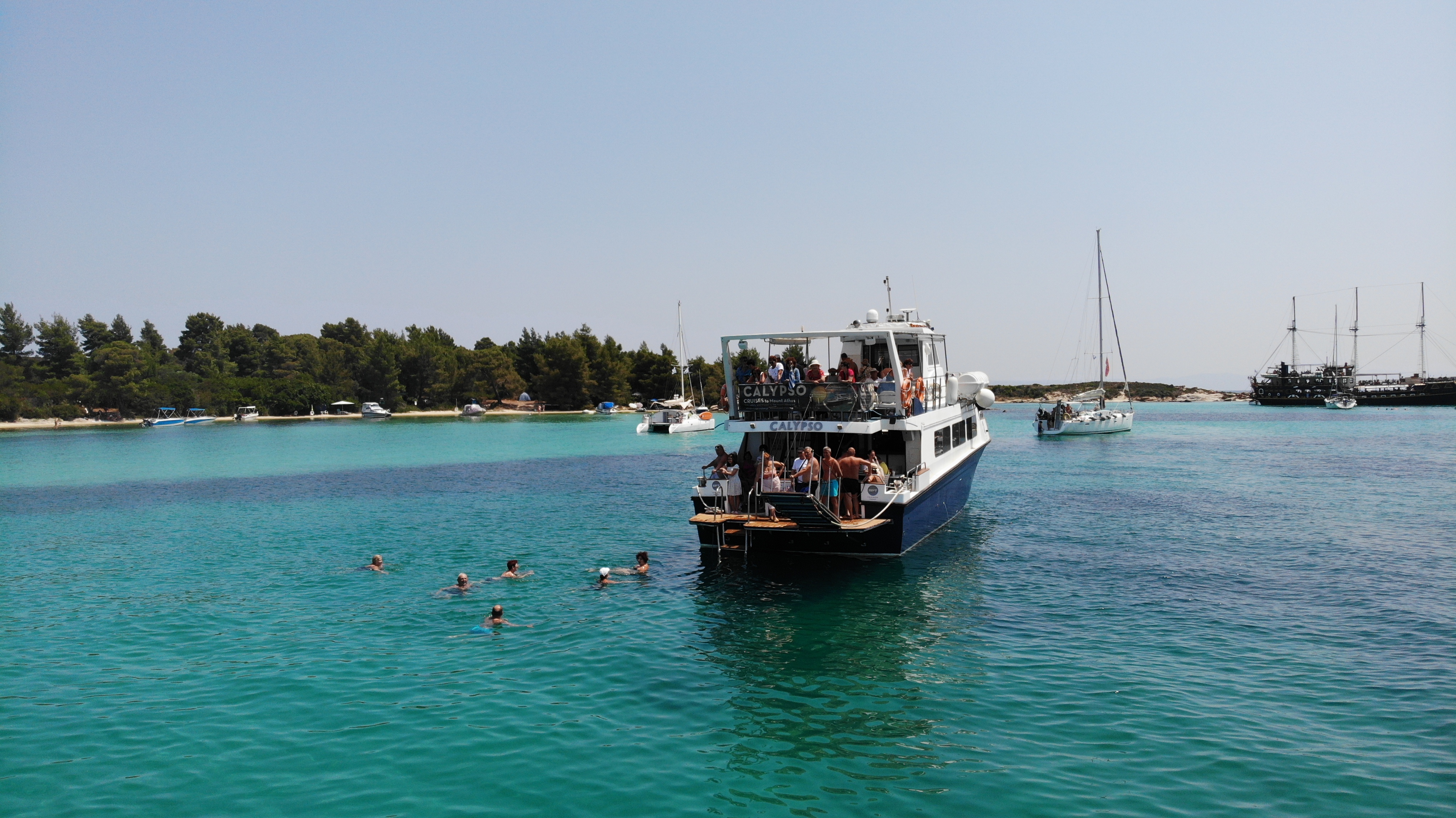 Eine Gruppe von Menschen schwimmt im türkisfarbenen Meer neben dem Boot von Calypso Cruises Ouranoupoli während einer Bootsfahrt von Ouranoupoli zur Blauen Lagune.