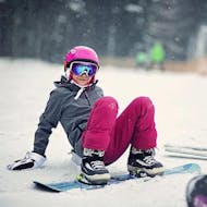 Ein Kind lächelt in die Kamera und sitzt im Schnee während ihrem Snowboardkurs (ab 6 J.) für Anfänger mit Feldberg Sports.