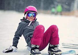 A kid smiling in the camera, sitting in the snow during her Snowboarding Lessons (from 6 y.) for First Timers from Feldberg Sports.