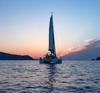 A catamaran with the colorful backdrop of the sunset during the Private Sunset Catamaran Trip along the Caldera with BBQ & Open Bar from Volcano Yachting Santorini.