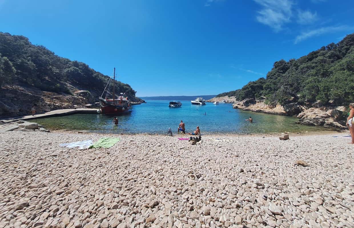 The M/B Kosljun Punat pirate ship is docked in a scenic cove on Plavnik Island, while visitors relax on the pebble beach and swim in the calm, turquoise sea.