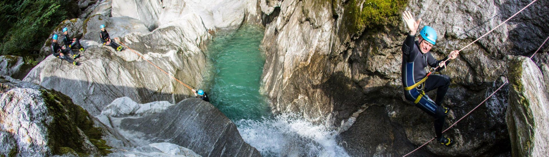 Ein Teilnehmer der Tour Canyoning “Blue Lagoon” für Mutige - Zemmschlucht von Mountain Sports Mayrhofen winkt in die Kamera während er sich in die Schlucht hinabseilt.