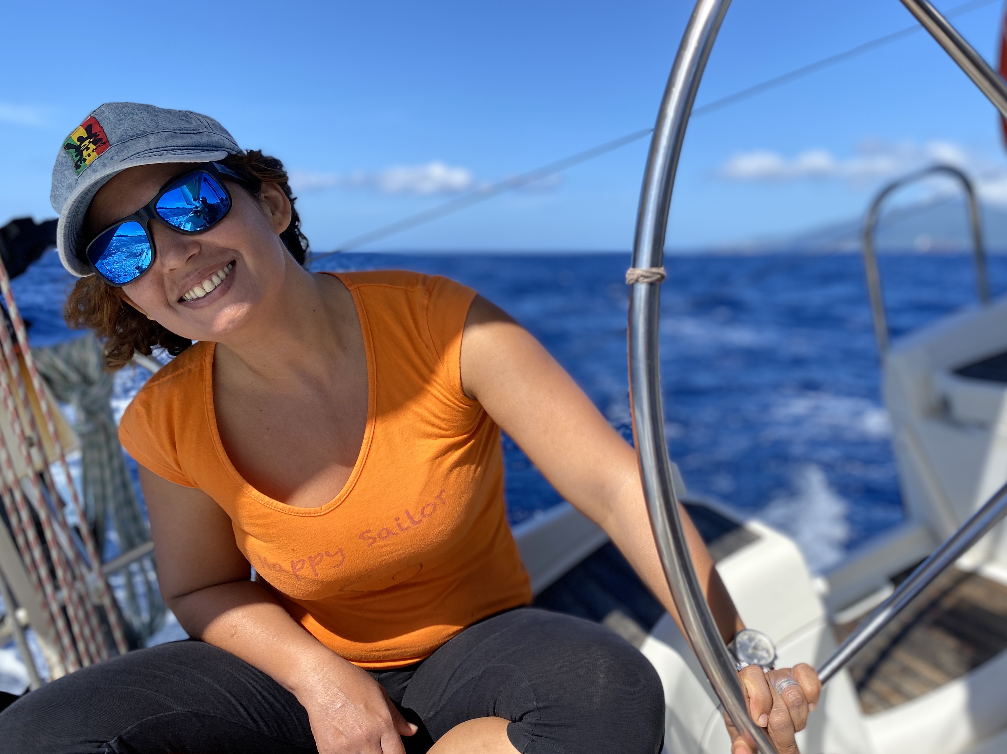 Eine lächelnde Frau mit Sonnenbrille entspannt sich am Steuer eines Segelboots auf dem offenen blauen Meer während eines Segeltörns nach Les Saintes mit Reve de Nav' Croisière.