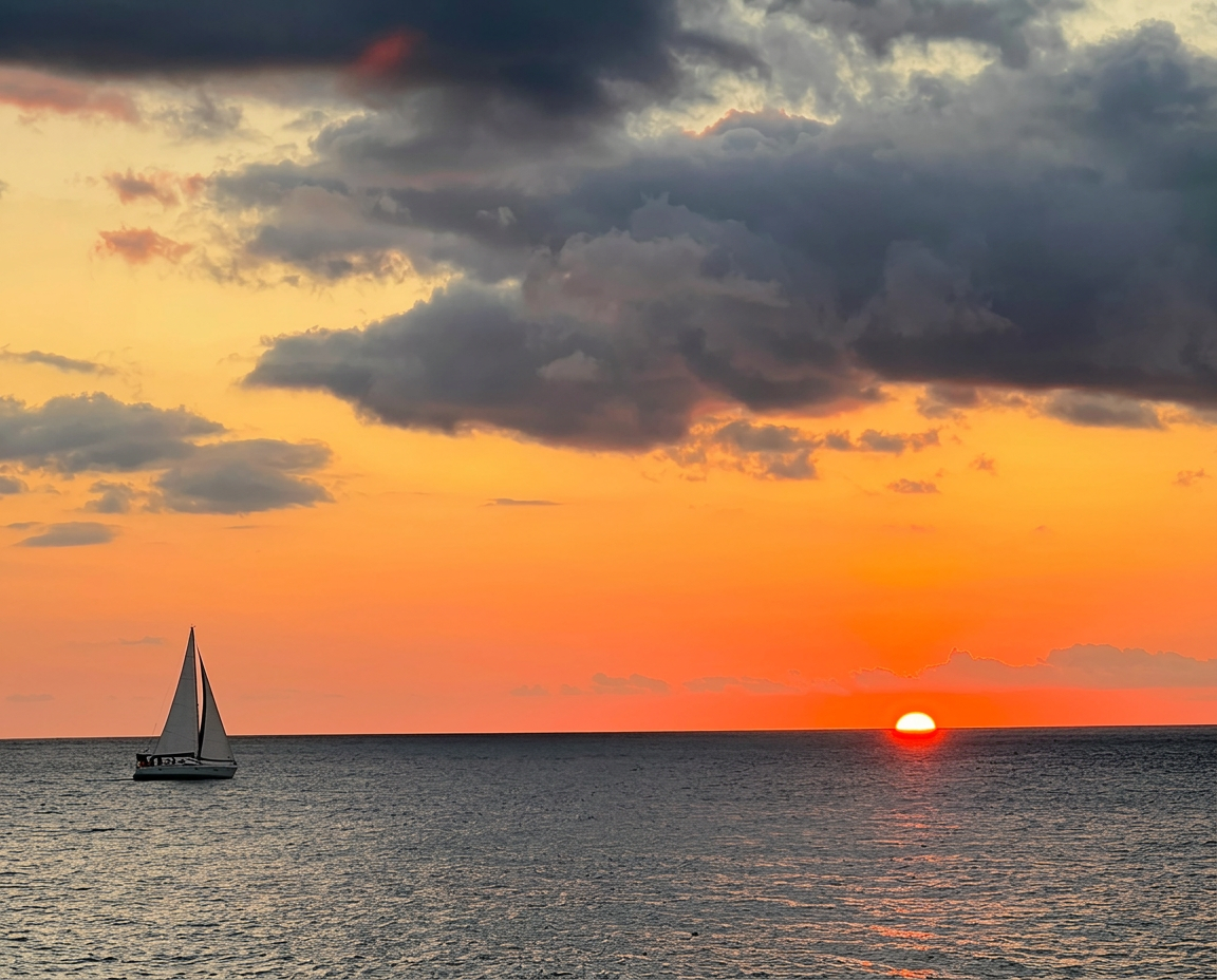 Ein Segelboot gleitet über die ruhige See vor einem leuchtend orangefarbenen Sonnenuntergang während eines Segeltörns nach Les Saintes mit Schnorcheln und Mittagessen von Reve de Nav' Croisière.
