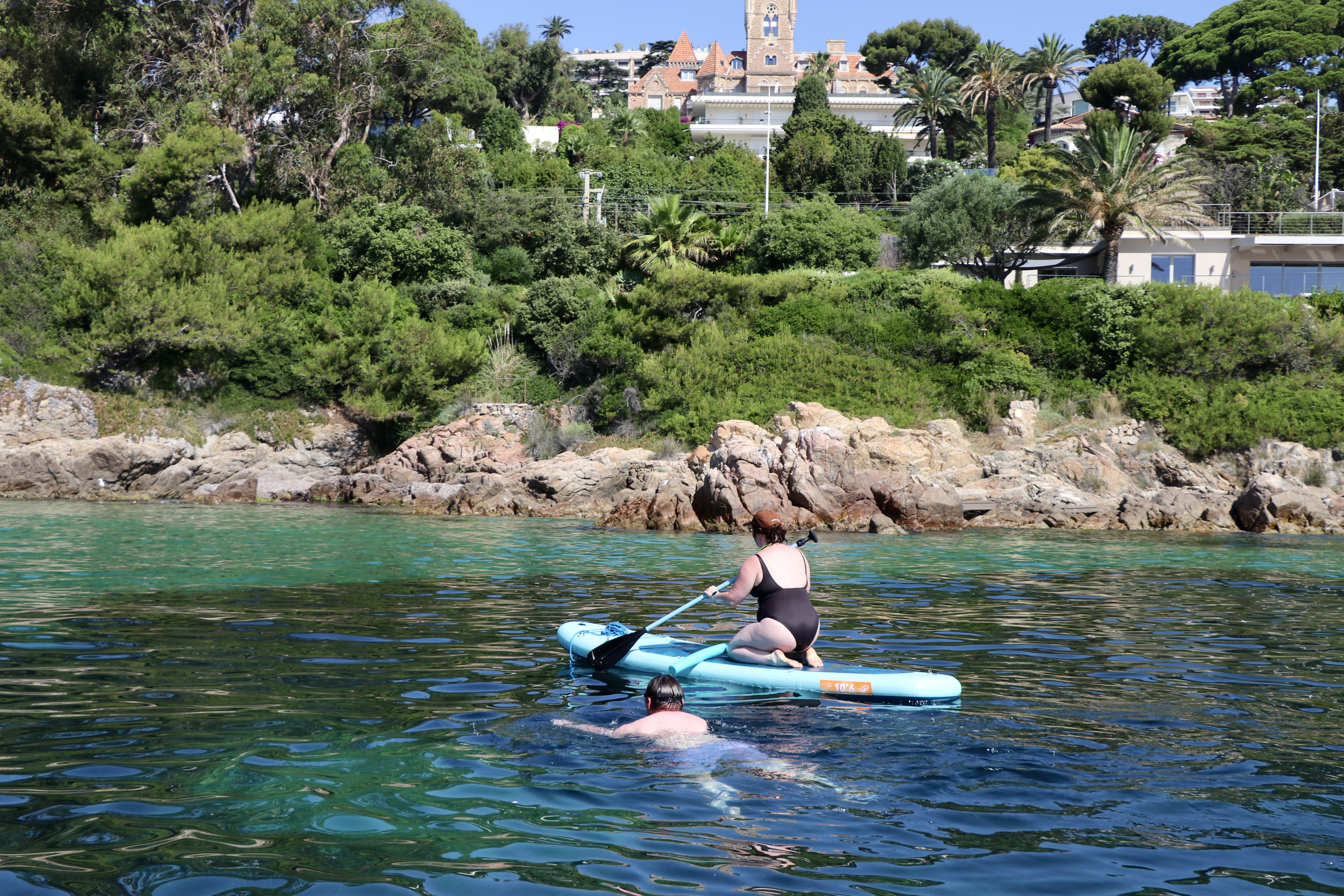 Pareja practicando paddle surf y nadando frente a la costa de Antibes. Un hombre nada mientras una mujer practica paddle surf en las aguas cristalinas durante una excursión en velero por Antibes con parada para nadar, organizada por Cap Sail Adventures Antibes.