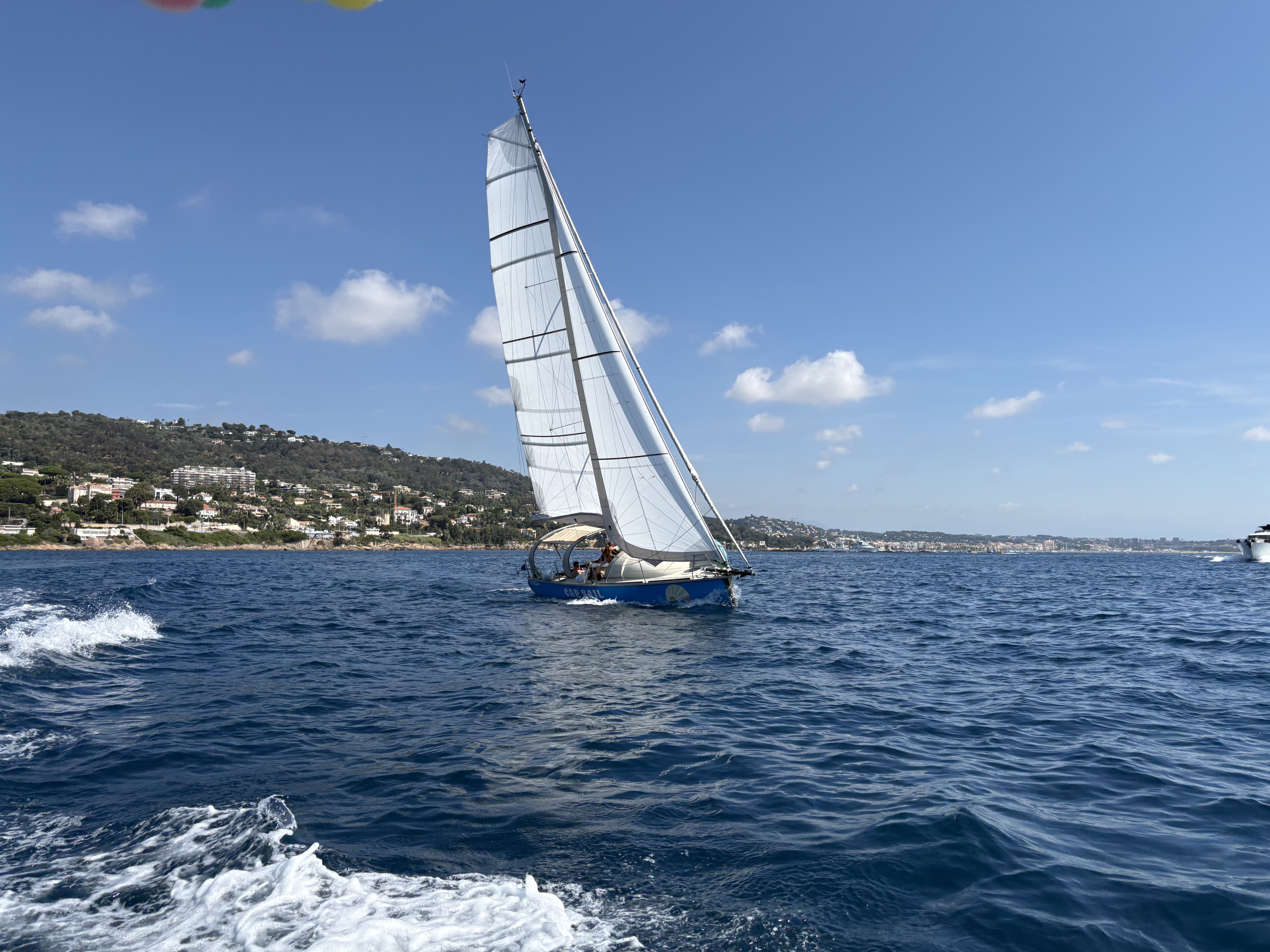 A blue and white sailboat from Cap Sail Aventures Antibes sails on the bright blue sea off the coast of Antibes during a sailing trip with breakfast and a stop for a swim.