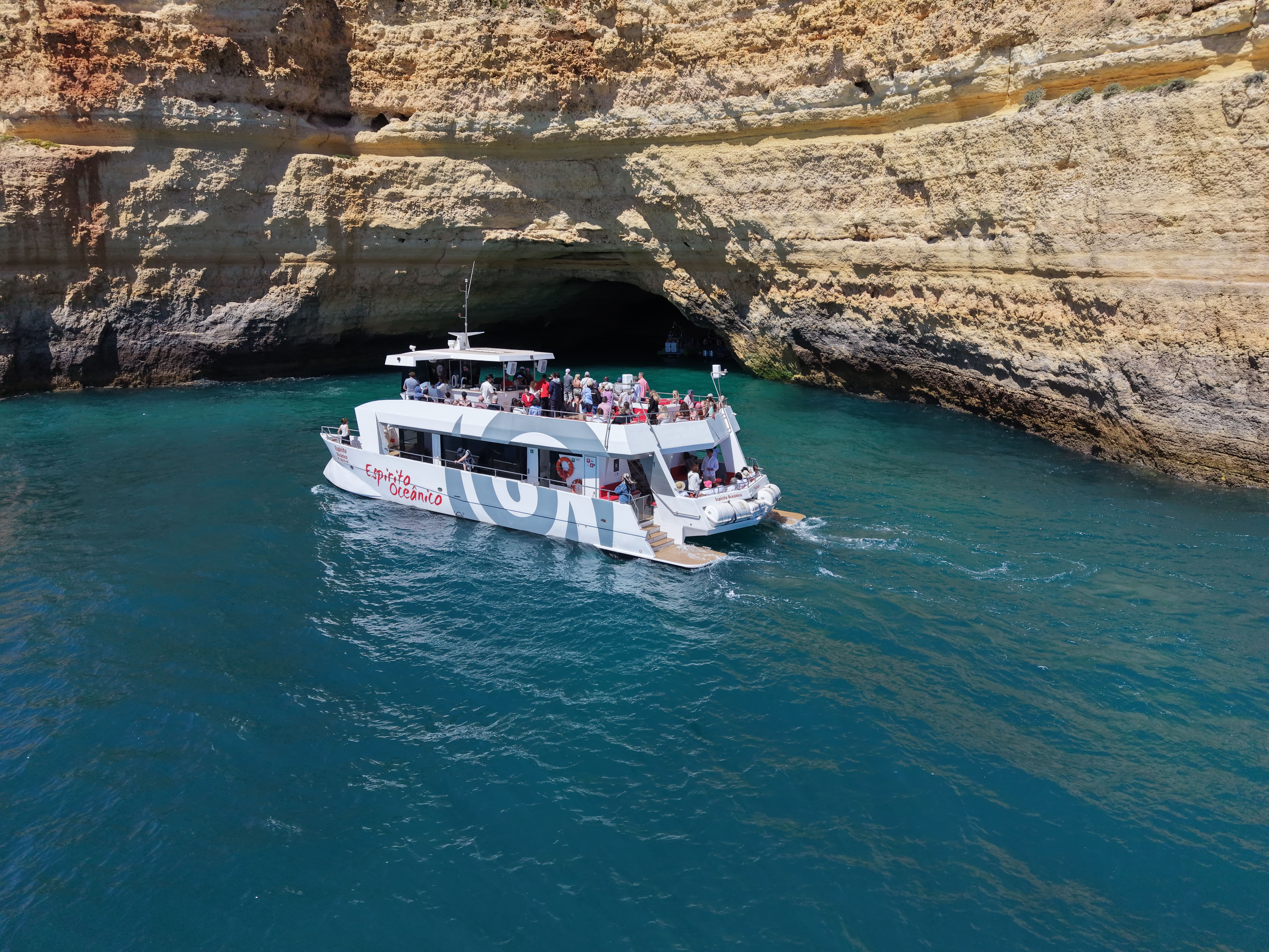 Una barca turistica bianca di AlgarExperience naviga con i passeggeri lungo le splendide scogliere e le grotte marine durante una crociera a Portimão: Grotte di Benagil con brunch e open bar.