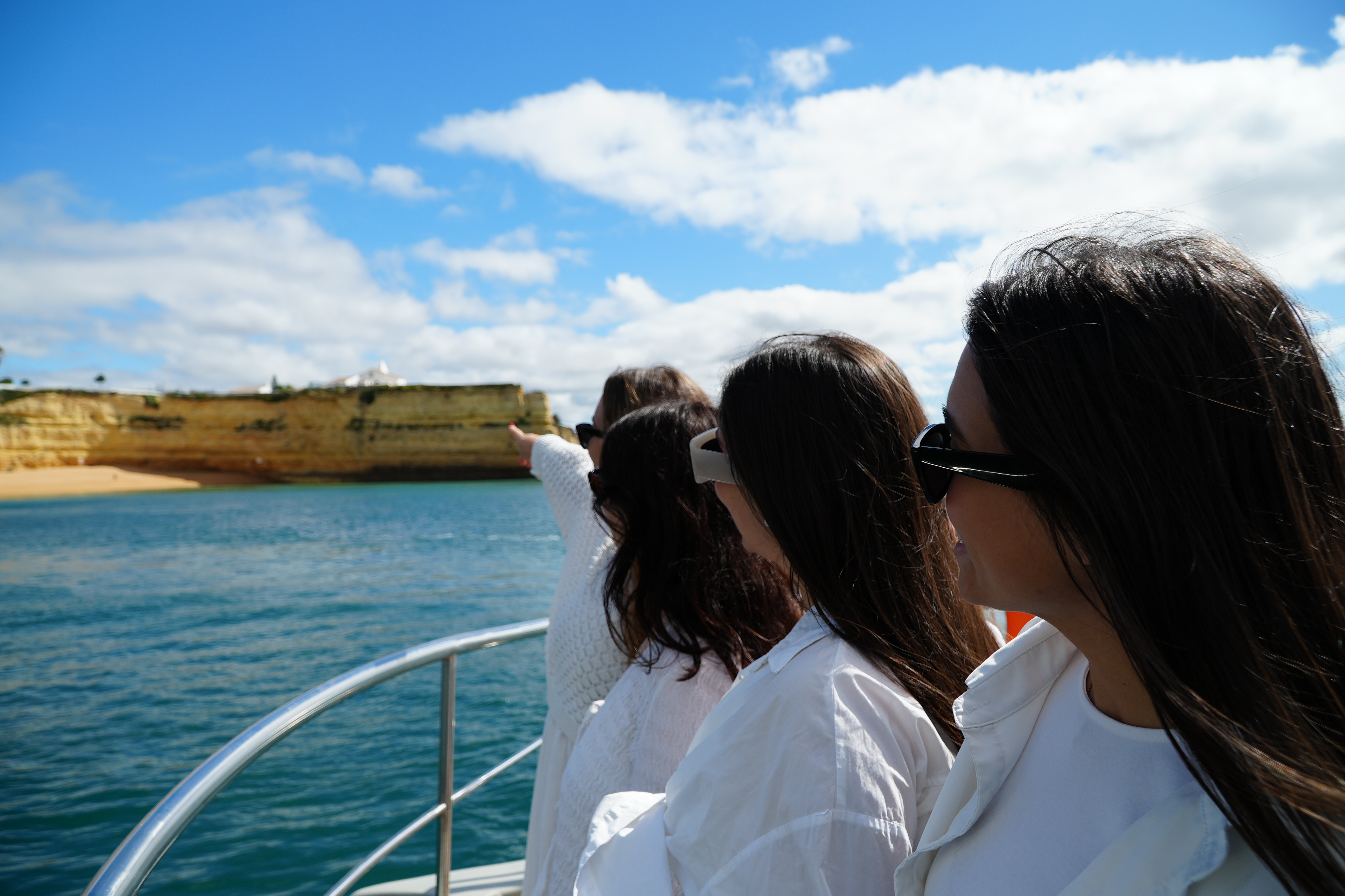 Four young women in white shirts admire the golden cliffs and blue sea from a boat during the Portimão - Benagil Caves & Coastline tour by AlgarExperience.
