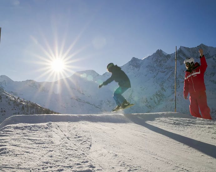 Ein Snowboarder springt unter der Aufsicht seines Snowboardlehrers beim Snowboardkurs für Erwachsene (ab 13 J.) für Anfänger.