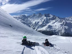 Twee snowboarders zitten op de piste tijdens hun Snowboardles voor volwassenen (vanaf 13 jaar) voor beginners van de Zwitserse Skischool Saas-Grund.