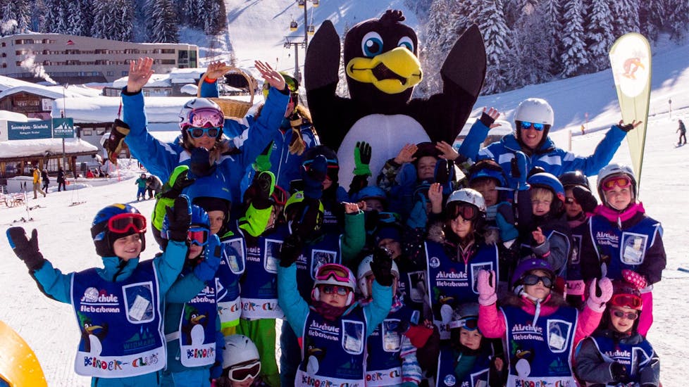 Eine große Gruppe Kinder winkt der Kamera gemeinsam mit ihrem Skilehrer entgegen während dem Kinder Skikurs "BOBOs Miniclub" (3-4 Jahre) - Anfänger der Skischule Fieberbrunn Widmann Mountain Sports.