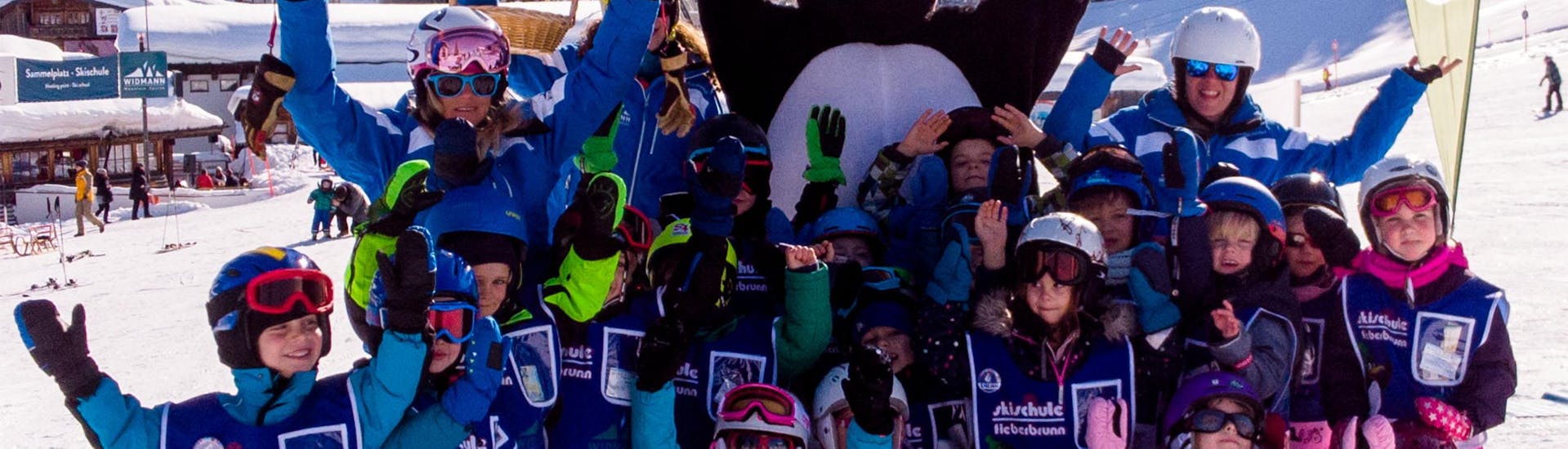 A large group of children wave to the camera together with their ski instructor during the kids ski lessons "BOBOs Miniclub" (3-4 years) - beginner of the Ski School Fieberbrunn Widmann Mountain Sports.