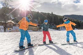 Two instructors from Skischule Thommi at Nassfeld are showing a child how to stand during its snowboarding lessons for beginners.