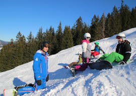 Des snowboarders assis sur la piste lors des cours de snowboard pour enfants (7-12 ans) pour tous les niveaux de l'école de ski et de snowboard Ostrachtal.