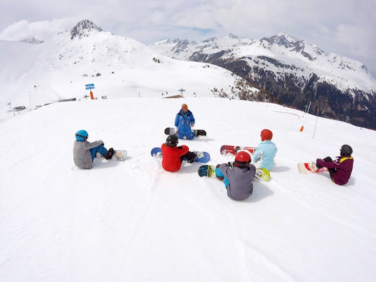 Tieners zitten in de sneeuw en luisteren naar hun snowboardleraar tijdens hun snowboardles voor kinderen en volwassenen - alle niveaus bij de skischool ESI Valfréjus.