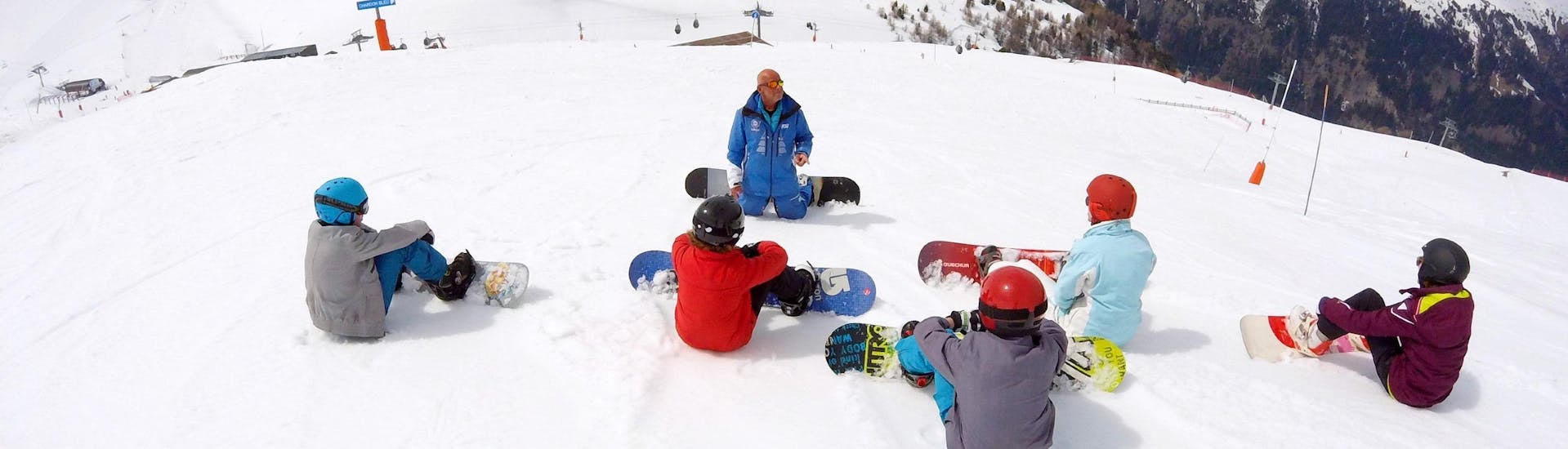 Teenagers are sitting in the snow and are listening to their snowboarding instructor during their Snowboarding Lessons for Kids & Adults - All Levels with the ski school ESI Valfréjus.