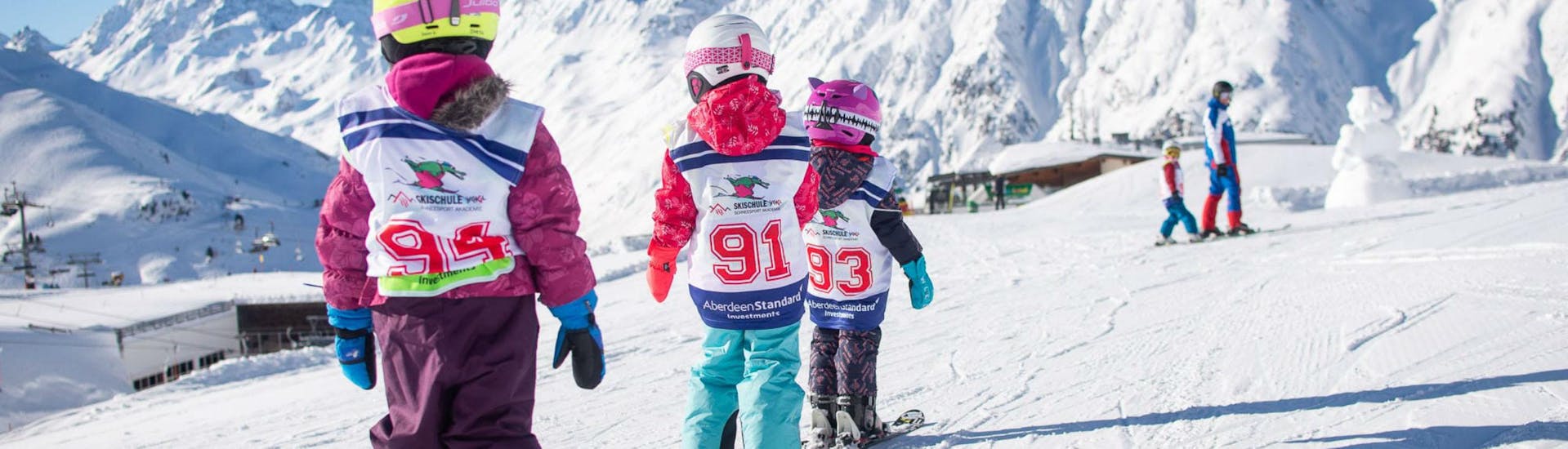 Three young kids are sking in a row behind one another along a flat plane of snow during their Kids Ski Lessons (6-15 years) - First Timer in the ski resort of Ischgl.