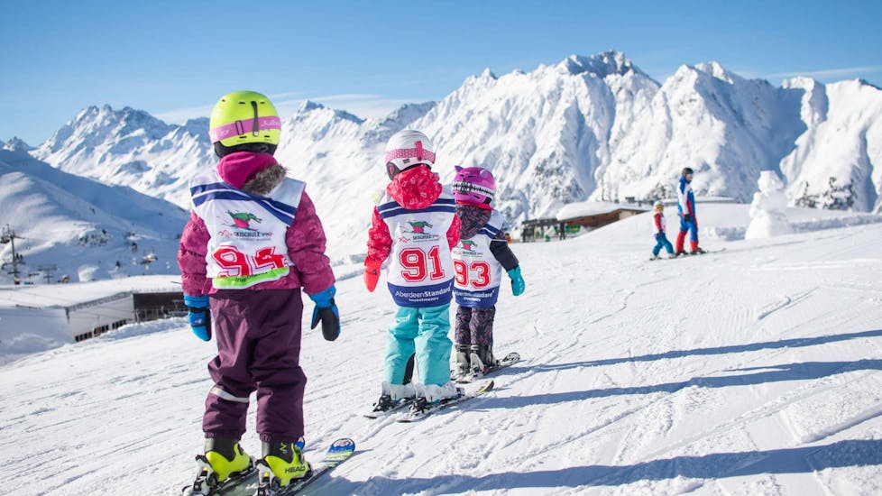 Three young kids are sking in a row behind one another along a flat plane of snow during their Kids Ski Lessons "Bambini" (3-5 years) - All Levels in the ski resort of Ischgl.