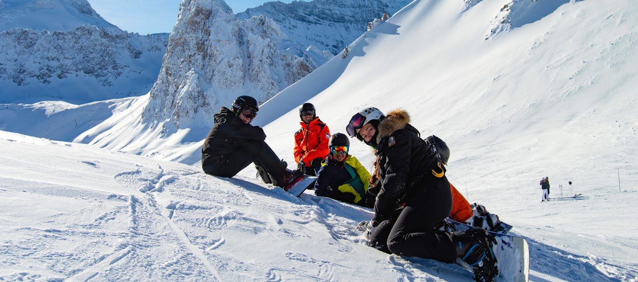 Die Snowboarder sitzen mitten auf dem Berg im Schnee, während sie mit Evolution 2 Val d'Isère Snowboardunterricht (ab 8 J.) für alle Levels nehmen.