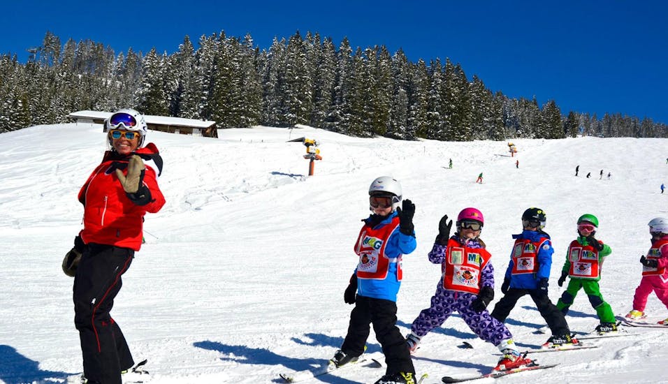 Een skileraar en haar groep blije kinderen zwaaien naar de camera terwijl ze oefenen met skiën tijdens de skilessen voor kinderen (vanaf 3 jaar) - alle niveaus met Skischule Kitzbühel Rote Teufel.