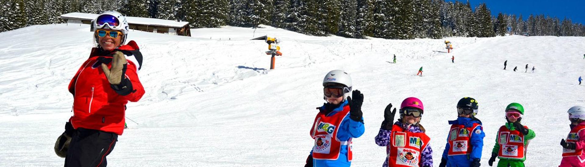 Eine Skilehrerin und ihre Gruppe fröhlicher Kinder winken beim Kinder Skikurs (ab 3 Jahren) - Alle Levels mit der Skischule Kitzbühel Rote Teufel in die Kamera.